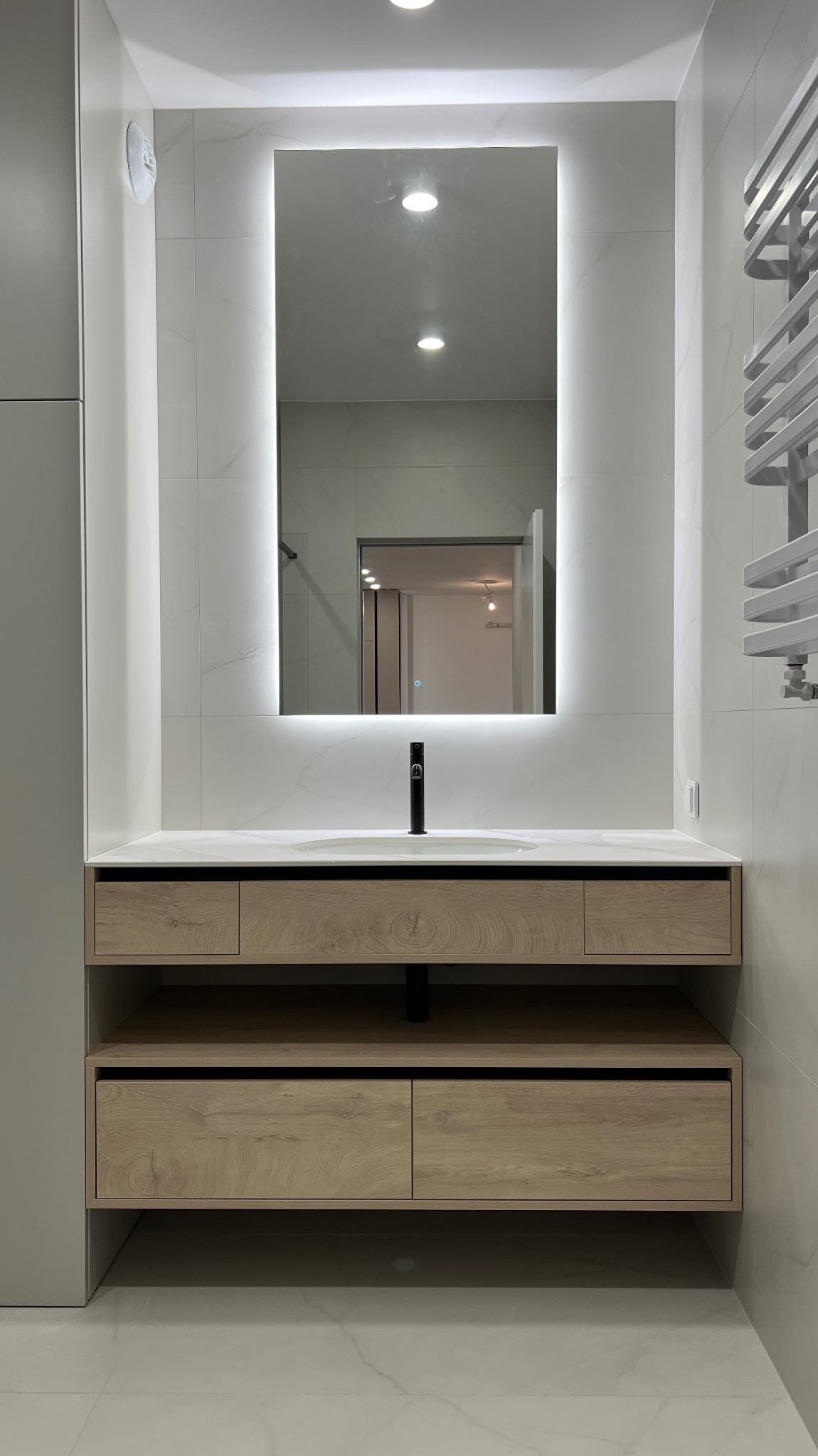 Nowoczesna szafka łazienkowa z drewnianymi frontami, umywalką i podświetlanym lustrem. Minimalistyczny design, płytki imitujące marmur, grzejnik drabinkowy.