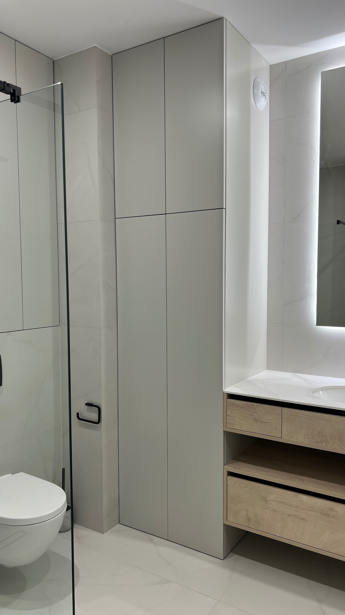 Jasna łazienka z zabudową meblową: szafa wnękowa z szarymi frontami i drewniana szafka pod umywalkę, białe płytki imitujące marmur, kabina prysznicowa.