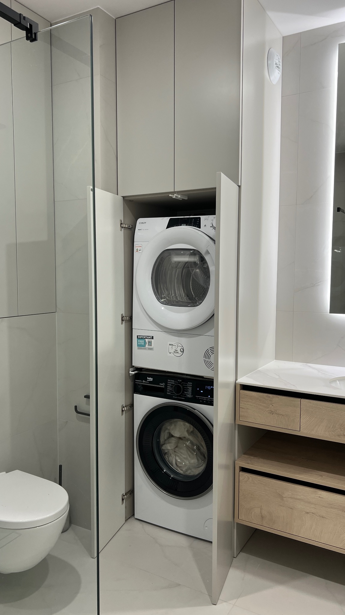 Biała pralka i suszarka w zabudowie meblowej łazienki, z otwartymi drzwiczkami szafy. Widoczna toaleta i fragment umywalki z szafką w stylu skandynawskim.