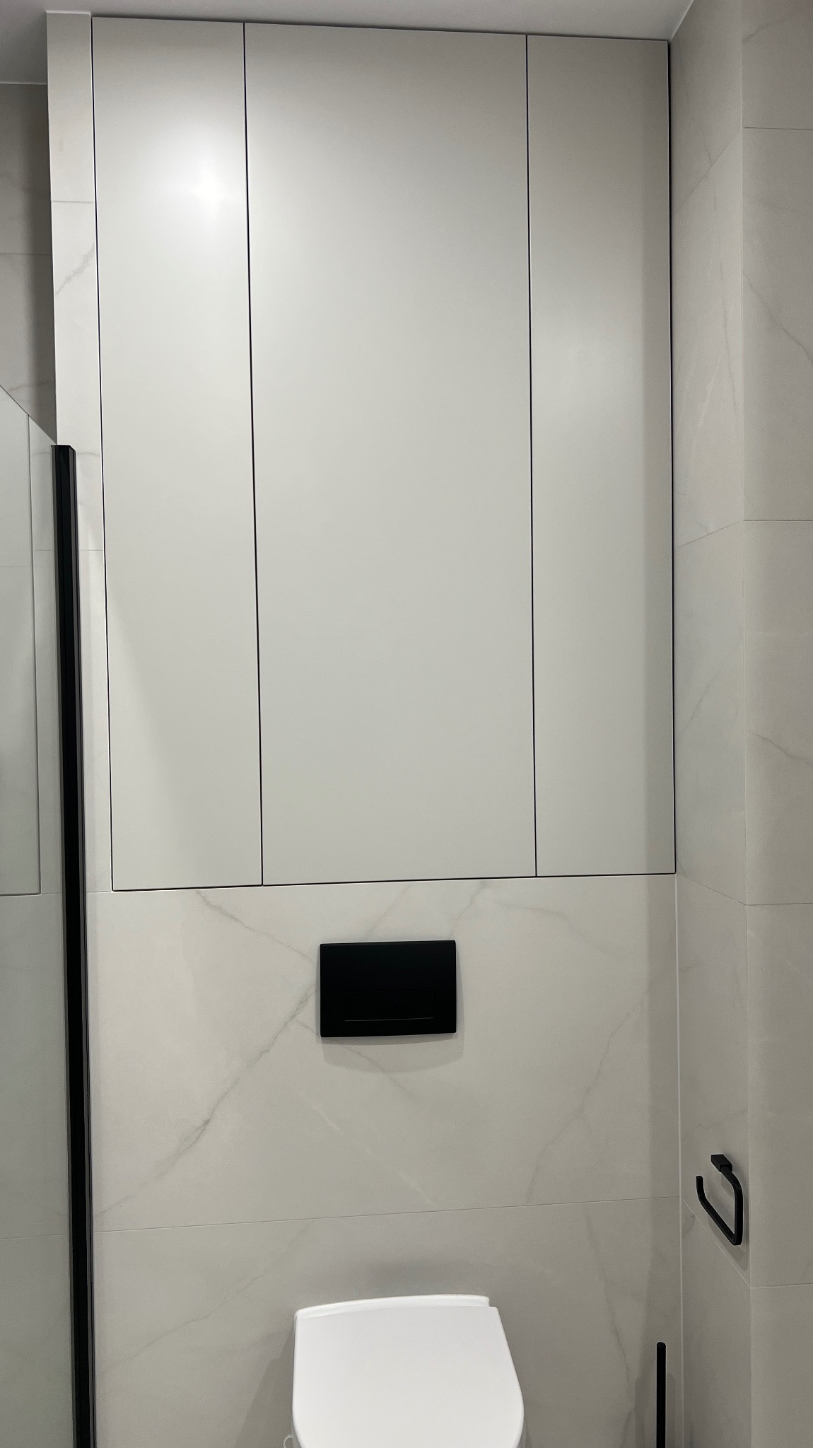 Białe szafki na wymiar w łazience nad toaletą, z czarnym przyciskiem spłuczki i marmurowymi płytkami na ścianie. Minimalistyczny design.