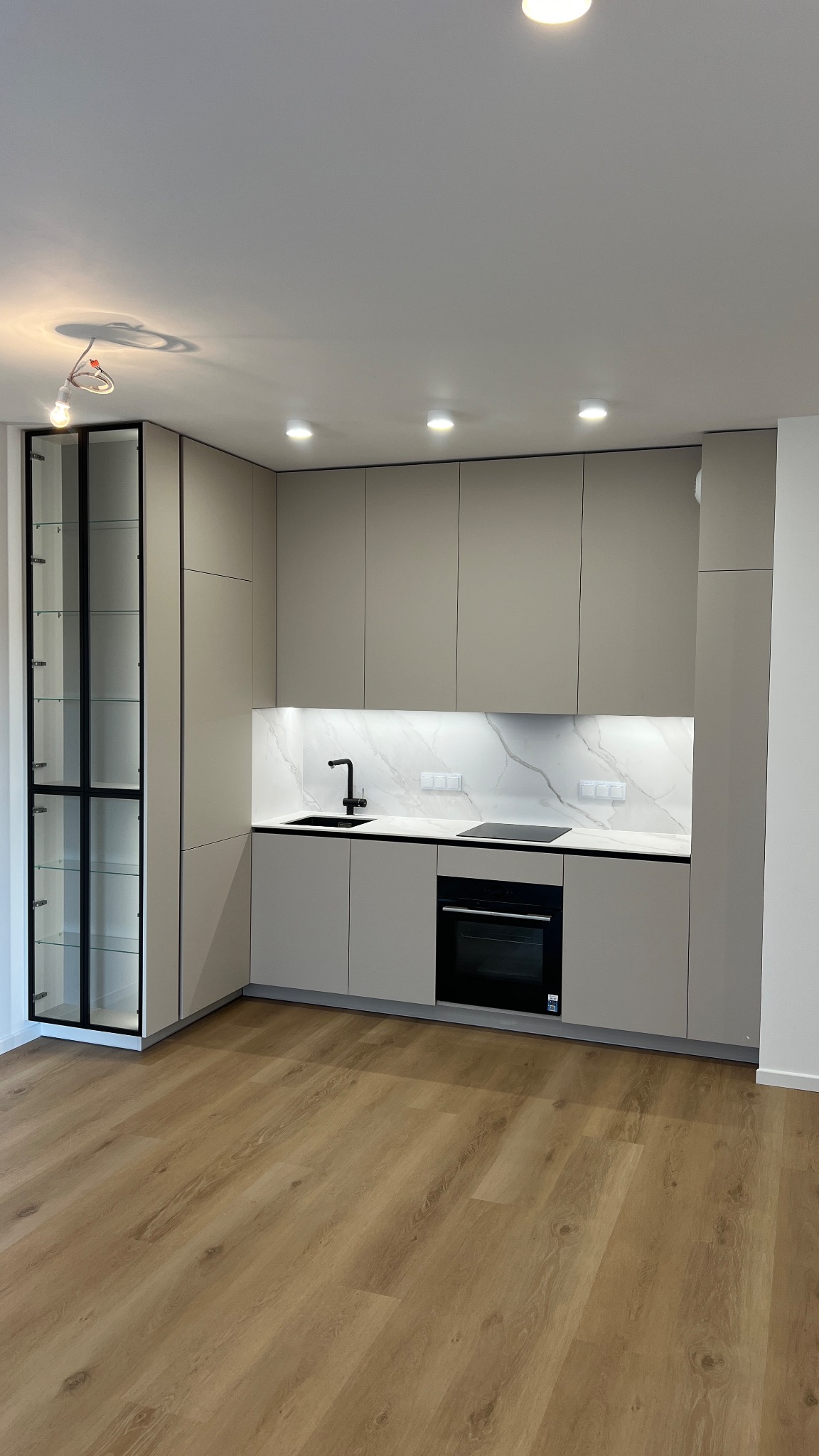 Jasna kuchnia na wymiar z czarnymi akcentami: szafka witrynowa, bateria i płyta indukcyjna. Minimalistyczny design, drewniana podłoga, nowoczesne oświetlenie.