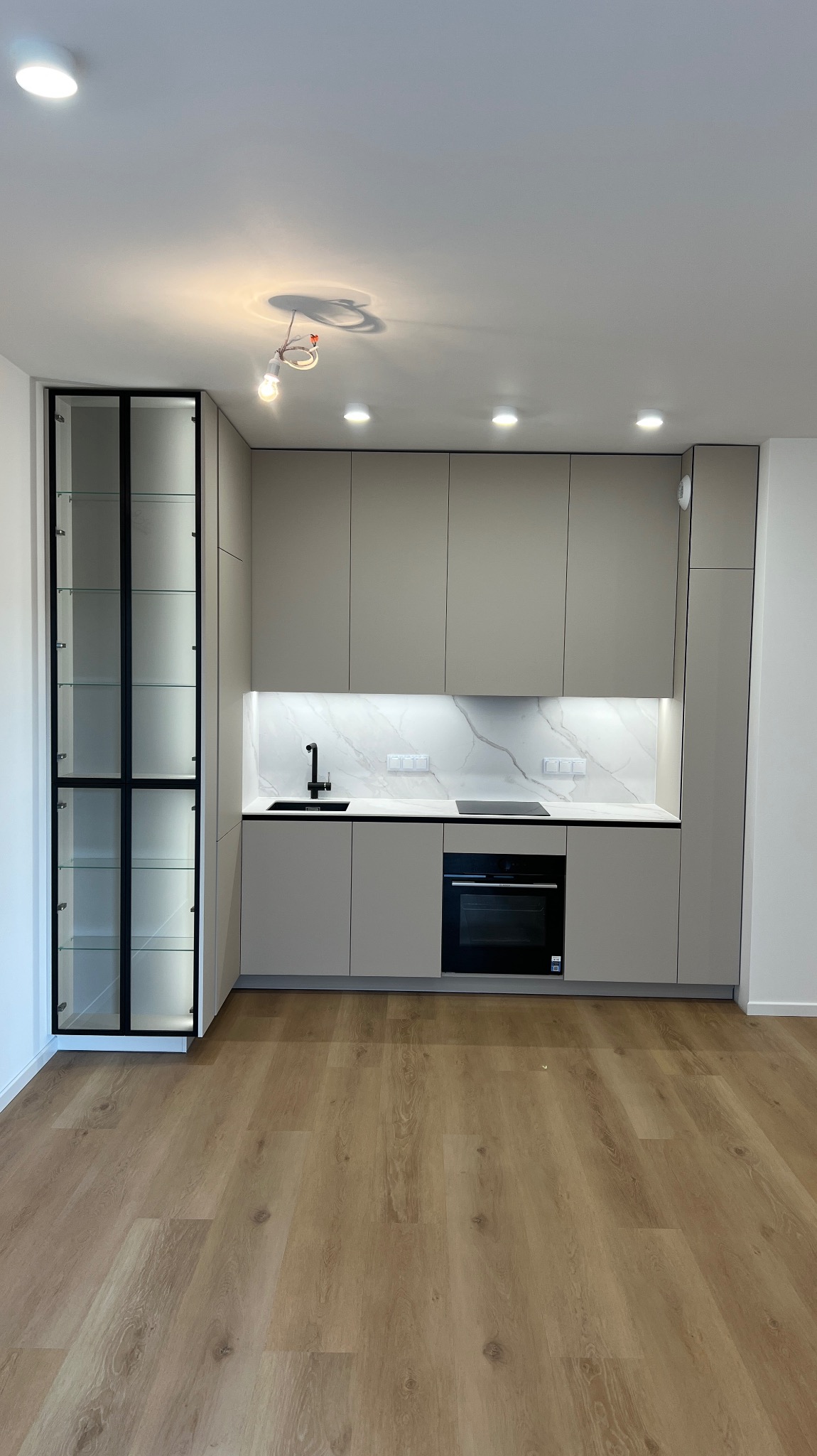 Nowoczesna kuchnia w odcieniach szarości z czarnymi akcentami, oświetleniem LED i drewnianą podłogą. Widoczna witryna z czarnymi ramami i szklanymi półkami.
