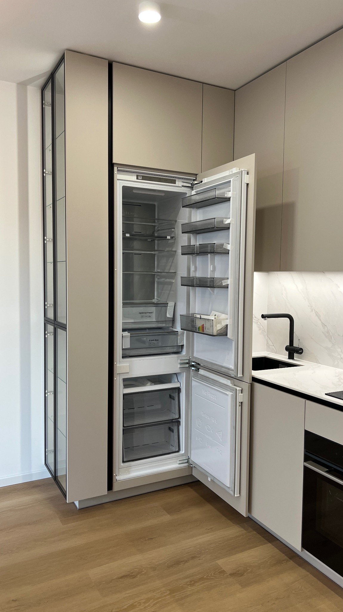 Beżowa kuchnia na wymiar z otwartą lodówką w zabudowie i szklaną witryną, drewniana podłoga, czarna bateria umywalkowa i marmurowy blat.