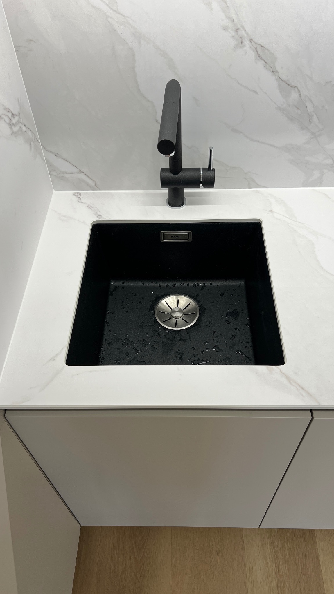 Nowoczesny zlew granitowy w czarnym kolorze, wkomponowany w biały blat z marmurowym wzorem. Bateria w matowej czerni. Ujęcie z bliska, podkreślające detale wykończenia.