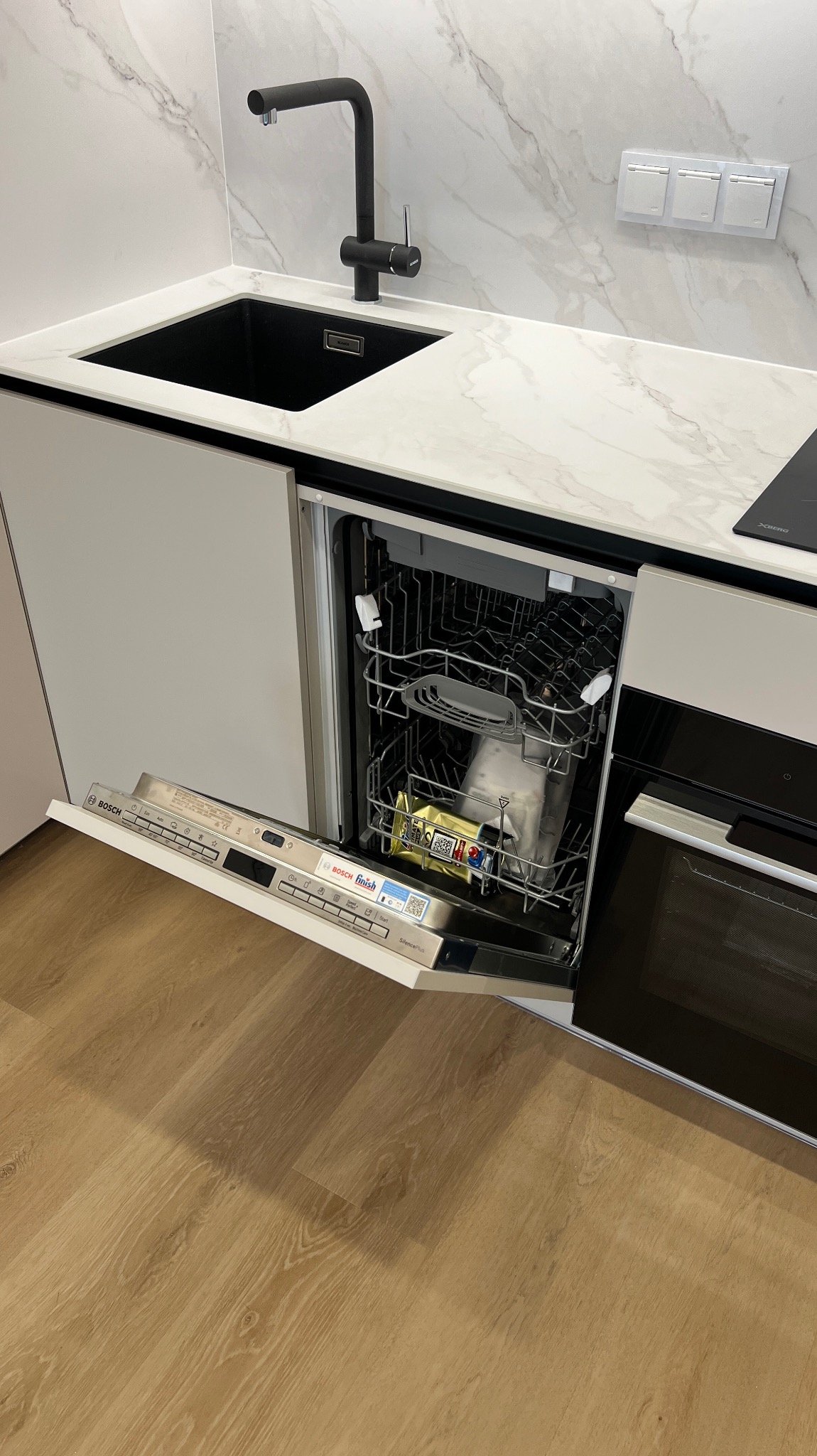 Nowoczesna kuchnia z otwartą zmywarką Bosch, czarną baterią i zlewozmywakiem, blat z marmurowym wzorem, drewnopodobna podłoga, minimalistyczny design.