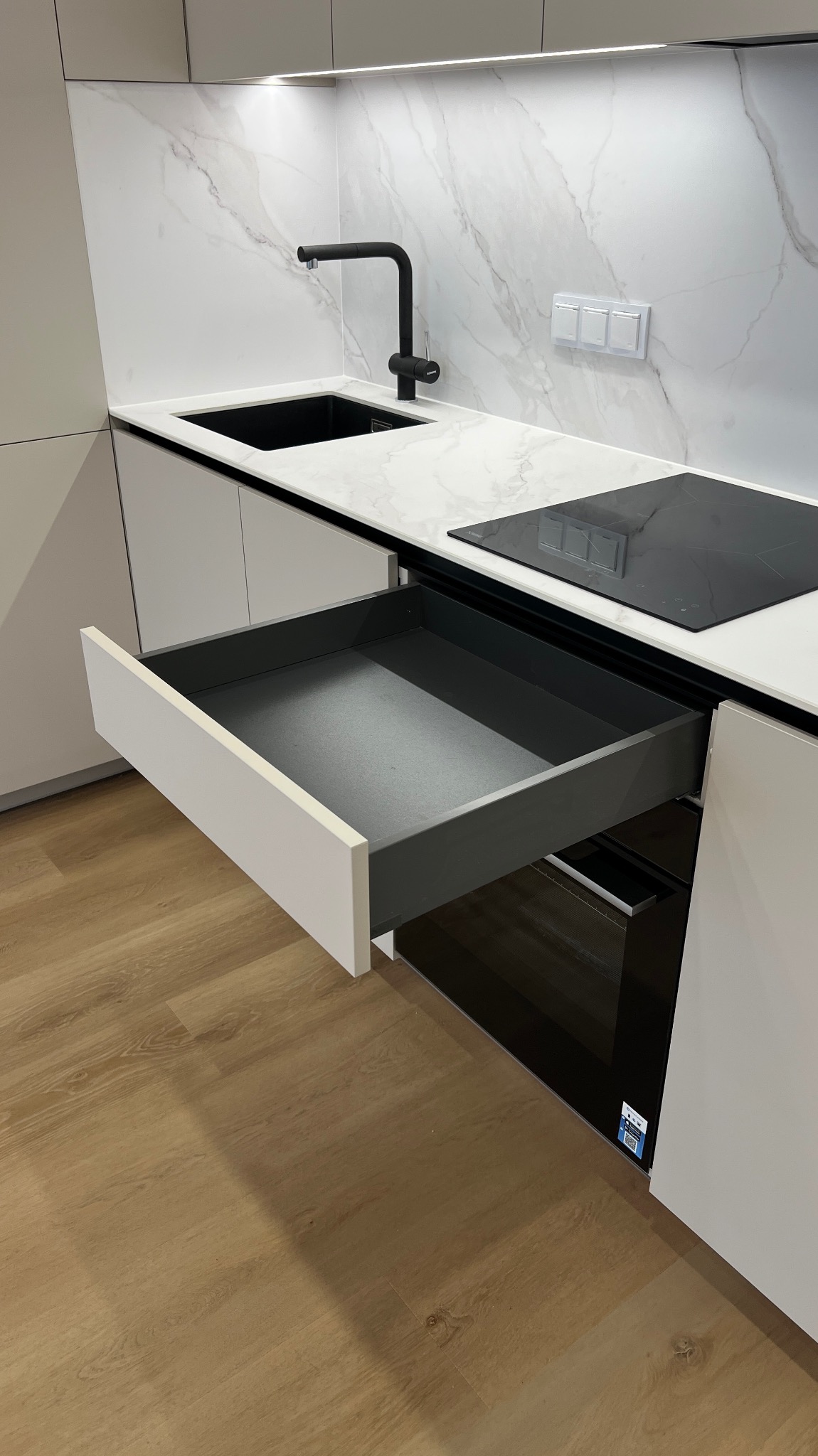 Nowoczesna kuchnia: białe fronty, marmurowy backsplash, czarna armatura i płyta indukcyjna. Otwarta szuflada z szarym wnętrzem. Jasna podłoga.