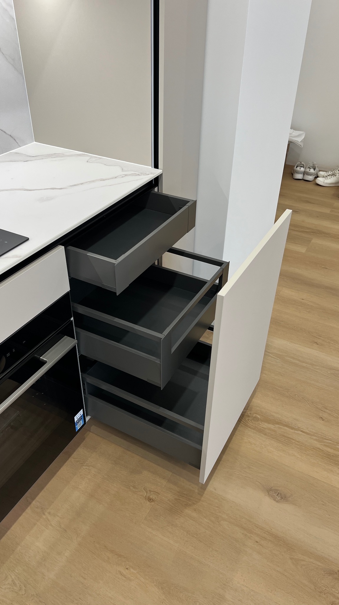 Nowoczesna kuchnia z szufladami cargo w kolorze grafitowym, wysuniętymi na zewnątrz. Minimalistyczny design, białe fronty i blat z wzorem marmuru.