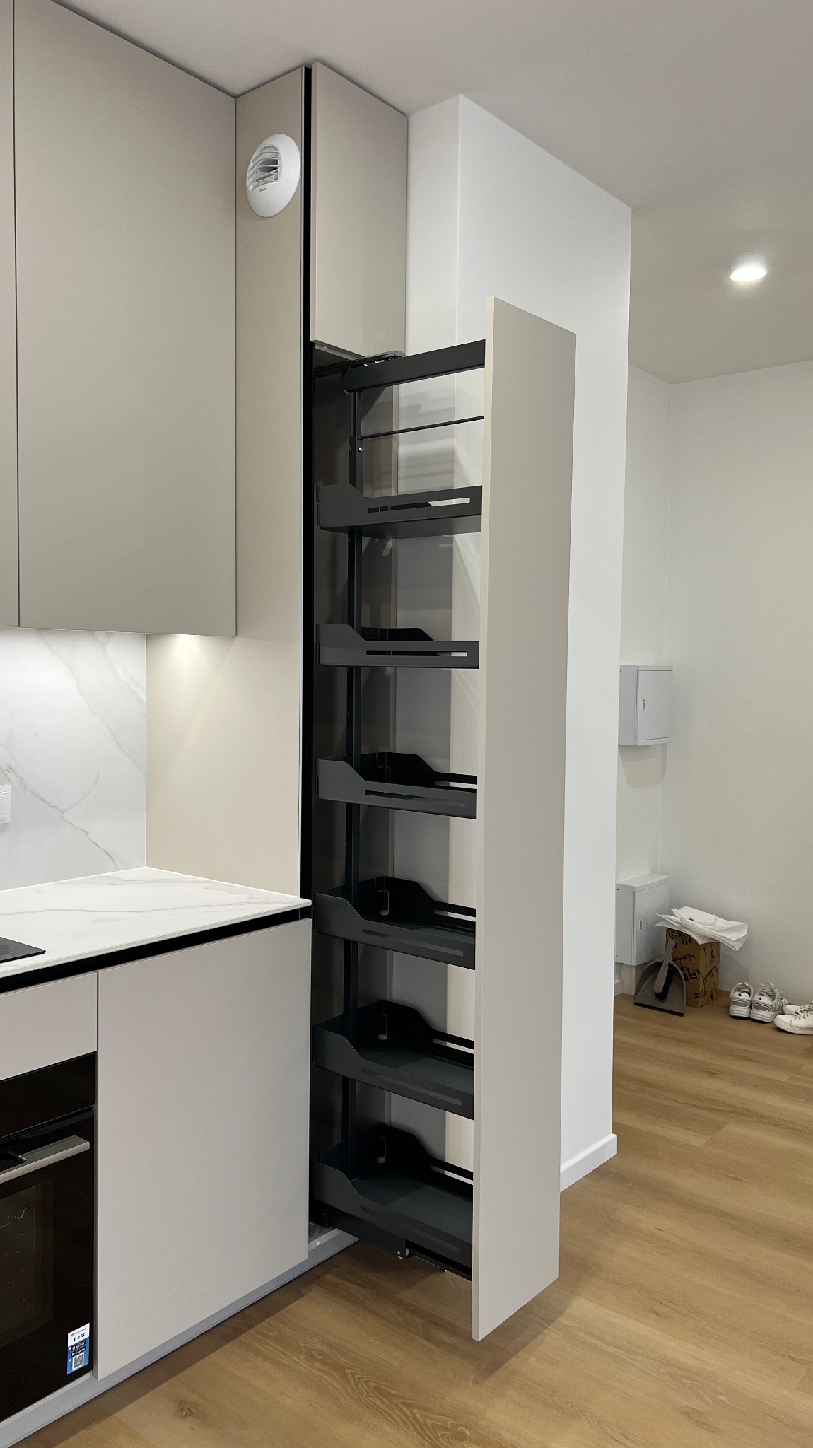 Nowoczesna kuchnia z wysuwaną szafką cargo w odcieniach szarości. Minimalistyczny design, jasne wnętrze, drewniana podłoga. Widoczny fragment blatu i piekarnika.