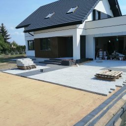 Krys bruk - Etap układania jasnoszarej kostki brukowej przed nowoczesnym domem z garażem. Częściowo utwardzony teren, paleta z kostką i przygotowane obrzeża.
