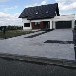 Krys bruk - Nowoczesny dom z podjazdem z szarej kostki brukowej, ułożonej we wzór z czarnymi akcentami. Widok na elewację domu i starannie wykończony teren.