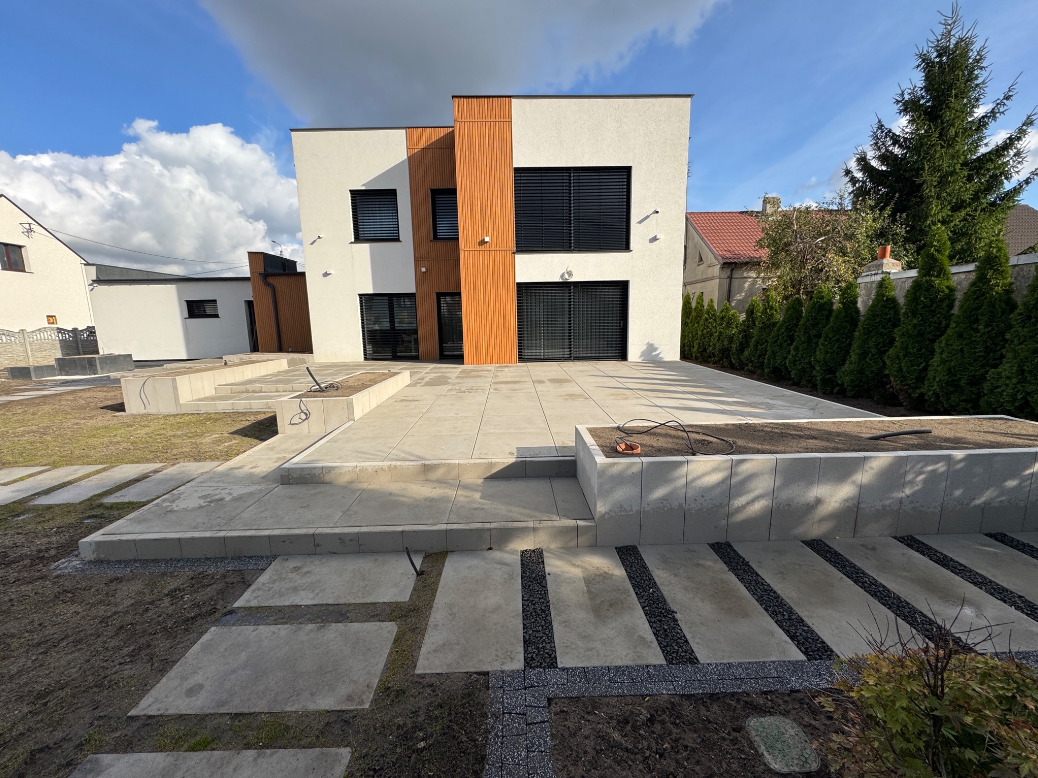 Nowoczesny dom z tarasem wyłożonym jasnoszarą kostką. Ścieżka z płyt prowadzi przez trawnik. Minimalistyczny design, geometryczne kształty, błękitne niebo w tle.