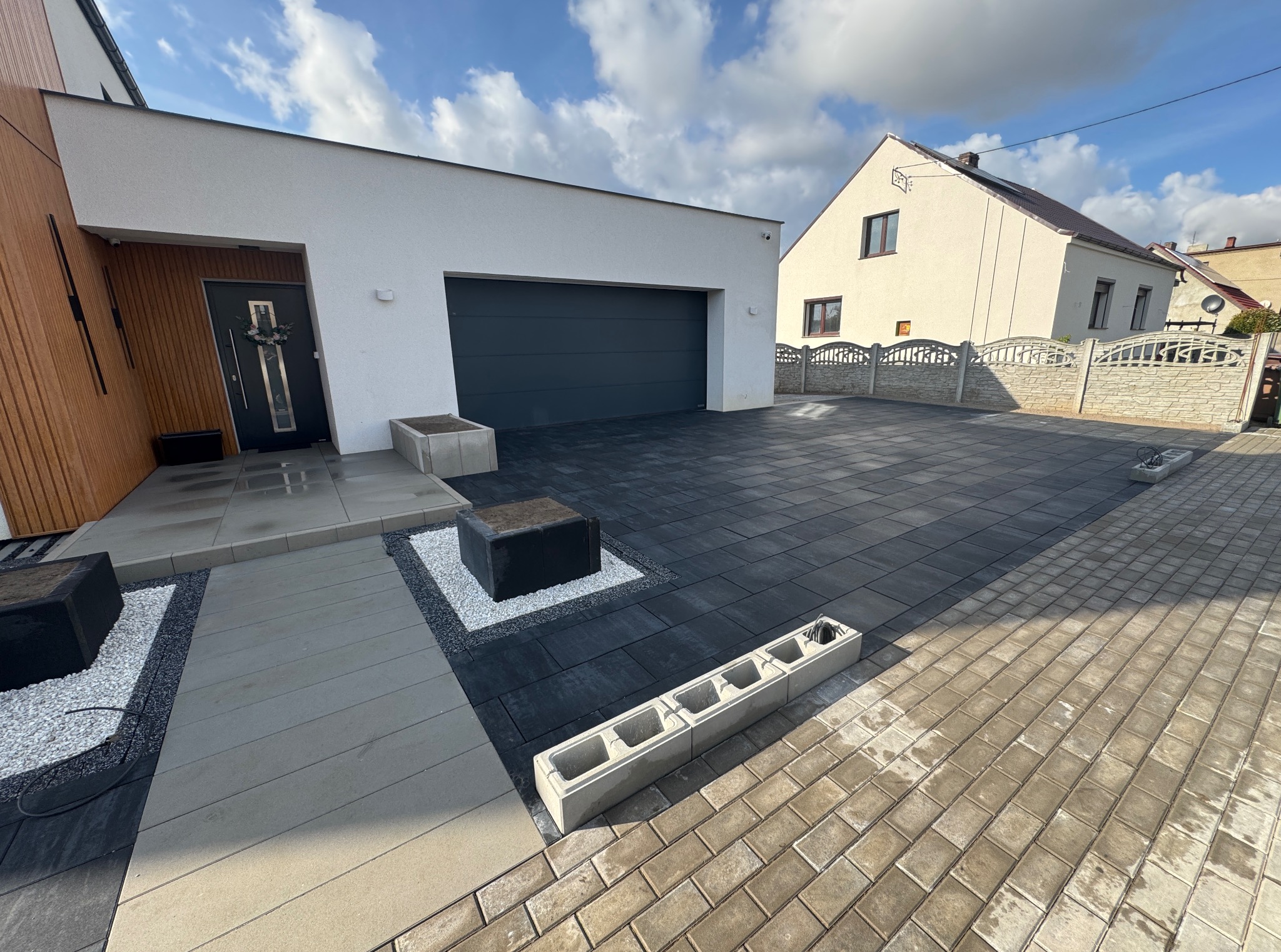 Nowoczesny podjazd z ciemnej kostki brukowej, jasne płyty chodnikowe i dekoracyjne elementy z białego kamienia. Dom z garażem w tle, słoneczny dzień.