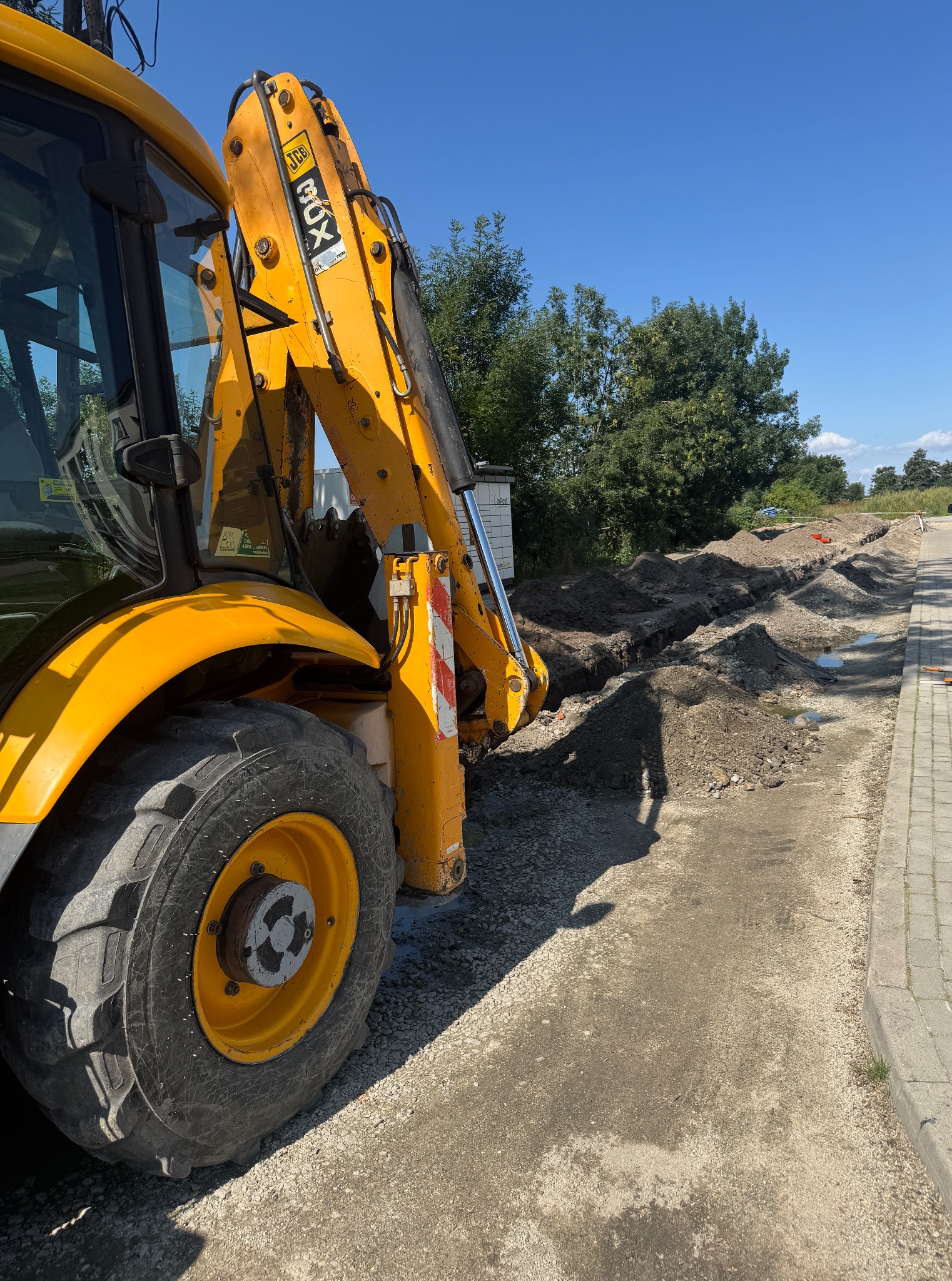 Żółta koparko-ładowarka JCB przy wykopie pod instalacje na żwirowej drodze, widok z boku, błękitne niebo w tle.