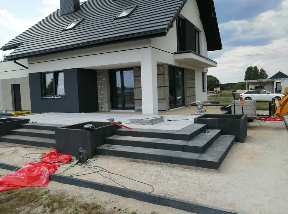 Nowoczesny dom z tarasem w trakcie budowy: schody z kostki brukowej, częściowo ułożona nawierzchnia, widoczne narzędzia i materiały budowlane. Jasne tło nieba.
