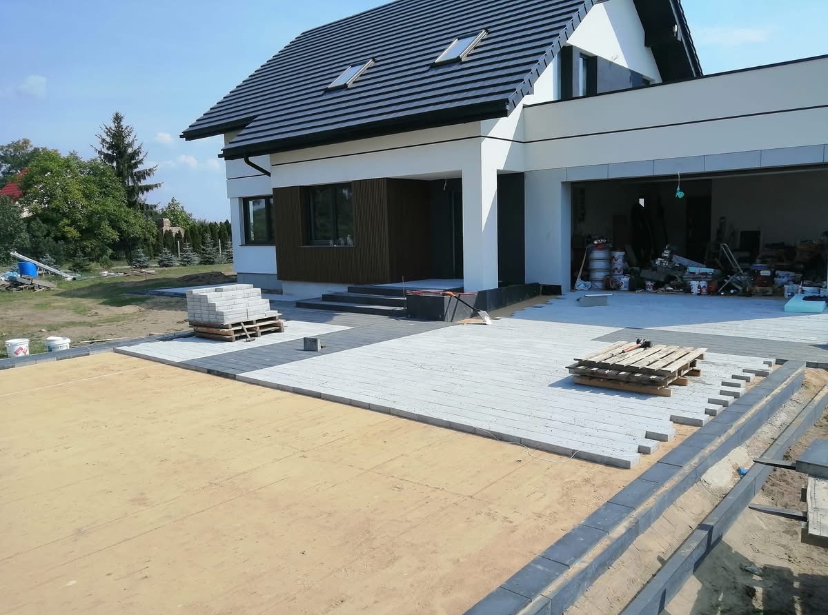 Etap układania jasnoszarej kostki brukowej przed nowoczesnym domem z garażem. Częściowo utwardzony teren, paleta z kostką i przygotowane obrzeża.