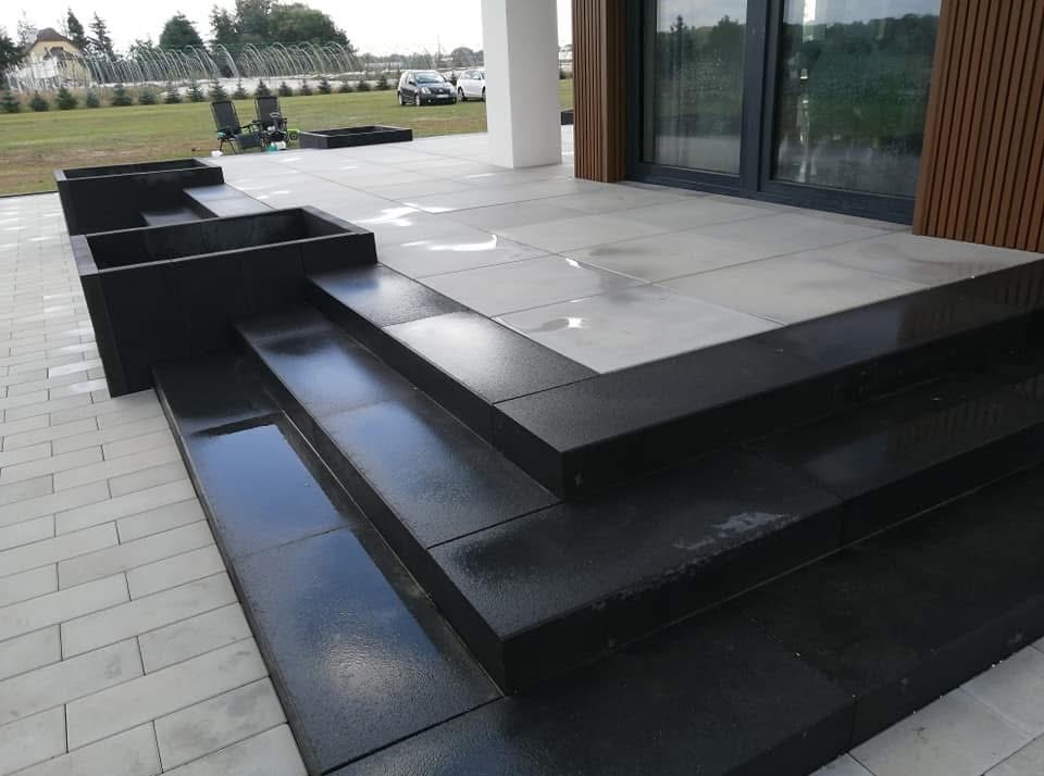 Nowoczesne schody z czarnej kostki granitowej prowadzące na taras z jasnoszarej kostki. Widoczne donice z kostki i trawnik w tle.