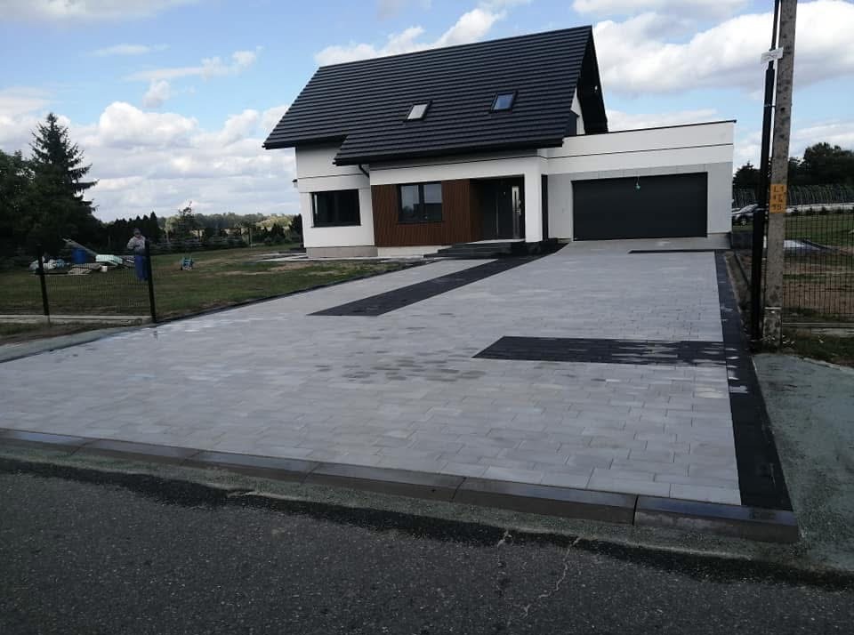 Nowoczesny dom z podjazdem z szarej kostki brukowej, ułożonej we wzór z czarnymi akcentami. Widok na elewację domu i starannie wykończony teren.