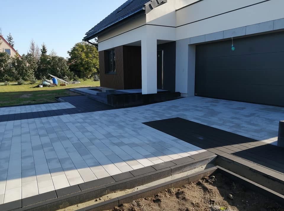 Nowoczesny podjazd z jasnoszarej kostki brukowej, urozmaicony ciemnymi wstawkami, prowadzący do domu z garażem. Widoczne obrzeża i fragment trawnika w tle.