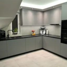 Szara, narożna kuchnia na wymiar z czarnym blatem i sprzętem AGD. Minimalistyczny design, oświetlenie podszafkowe, widok na schody z oknem.