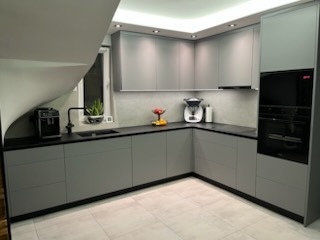 Szara, narożna kuchnia na wymiar z czarnym blatem i sprzętem AGD. Minimalistyczny design, oświetlenie podszafkowe, widok na schody z oknem.