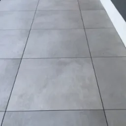 Szare, kwadratowe płyty tarasowe, ułożone równo na zewnątrz budynku. Widoczna krawędź ściany z białą listwą przypodłogową. Minimalistyczny design.