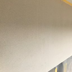RAFA - Szara, ziarnista faktura ściany wewnętrznej po nałożeniu tynku dekoracyjnego. Folia zabezpieczająca podłogę. Widoczna taśma malarska na górze ściany.