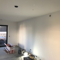 RAFA - Wnętrze w trakcie prac wykończeniowych: pakiety paneli podłogowych, narzędzia oświetlone lampą, świeżo pomalowane ściany i widok na zewnątrz przez okno.