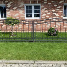 RAFA - Metalowe ogrodzenie posesji z dekoracyjnymi elementami na tle ceglanego domu z trzema oknami, trawnik i roślinność wzdłuż ogrodzenia.
