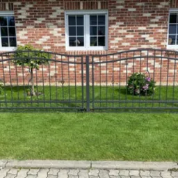 Metalowe ogrodzenie posesji z dekoracyjnymi elementami na tle ceglanego domu z trzema oknami, trawnik i roślinność wzdłuż ogrodzenia.