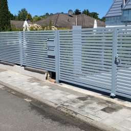 RAFA - Nowoczesne, szare ogrodzenie panelowe z poziomymi lamelami, z bramką wejściową, na tle zieleni i domów. Widok z poziomu chodnika, słoneczny dzień.