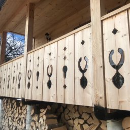 RAFA - Drewniana pergola z dekoracyjnymi wycięciami, oświetlona lampą. Pod konstrukcją składowane drewno opałowe. Widok na zimowy krajobraz.