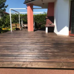 Ciemnobrązowy taras z desek w Soblówce, zadaszony pergolą, z grillem z cegły. Widok na ogród i dom.