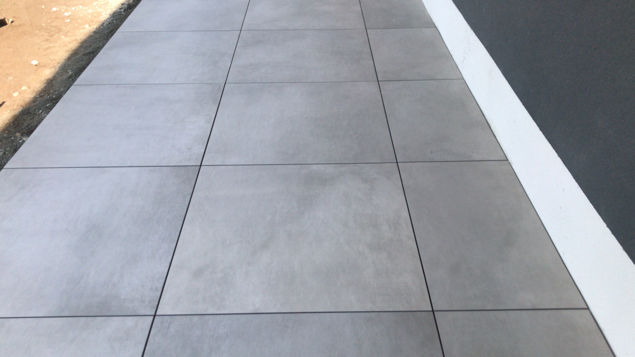 Szare, kwadratowe płyty tarasowe, ułożone równo na zewnątrz budynku. Widoczna krawędź ściany z białą listwą przypodłogową. Minimalistyczny design.