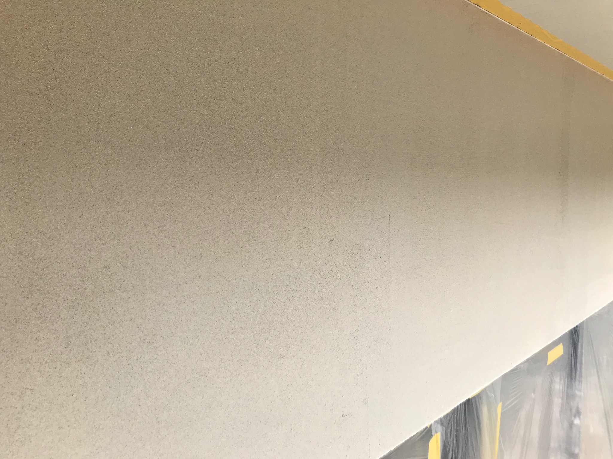 Szara, ziarnista faktura ściany wewnętrznej po nałożeniu tynku dekoracyjnego. Folia zabezpieczająca podłogę. Widoczna taśma malarska na górze ściany.