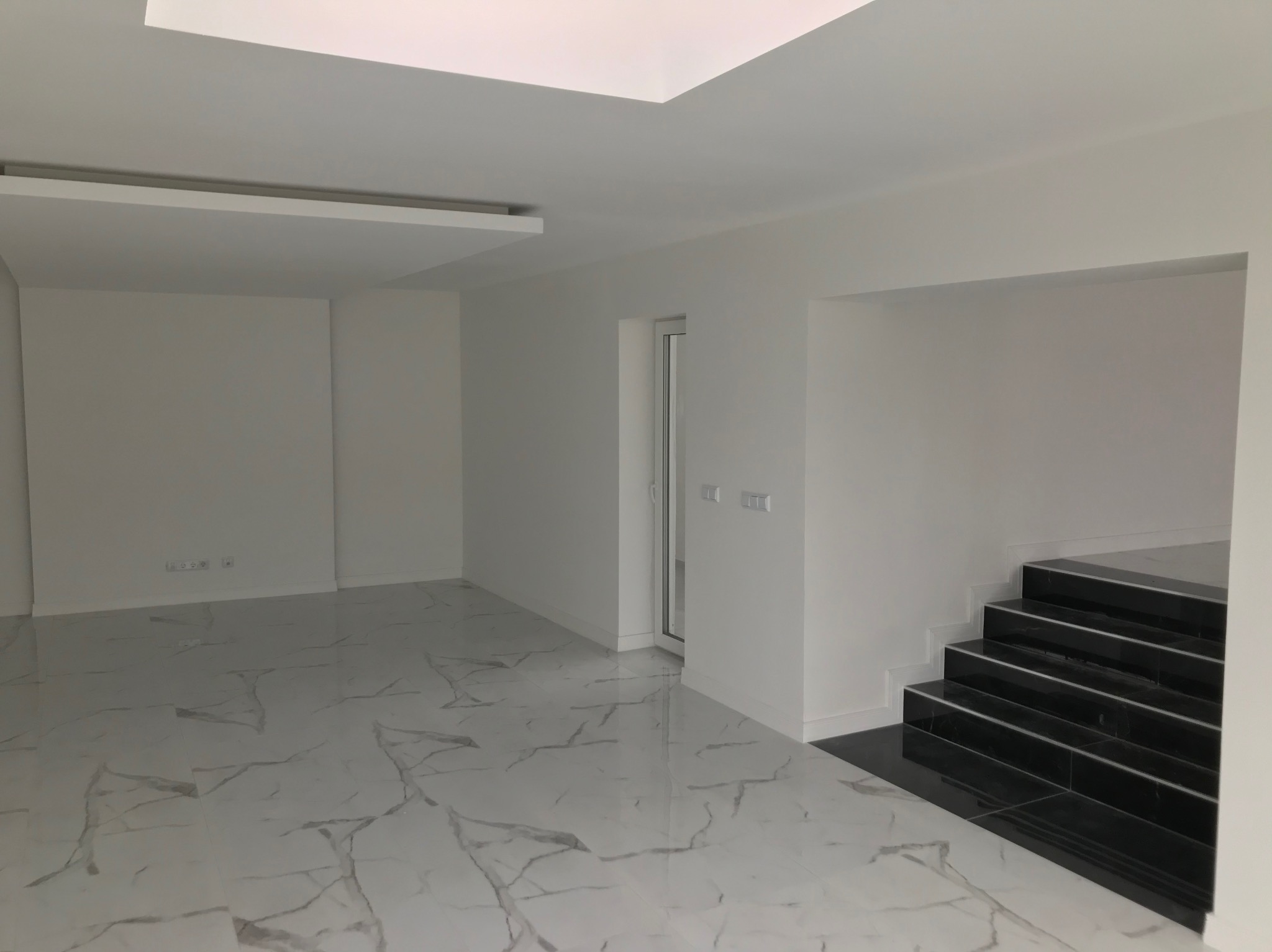 Nowoczesne wnętrze z białymi ścianami, marmurową podłogą i czarnymi schodami. Widoczne detale wykończenia sufitu i minimalistyczne gniazdka elektryczne przy podłodze.