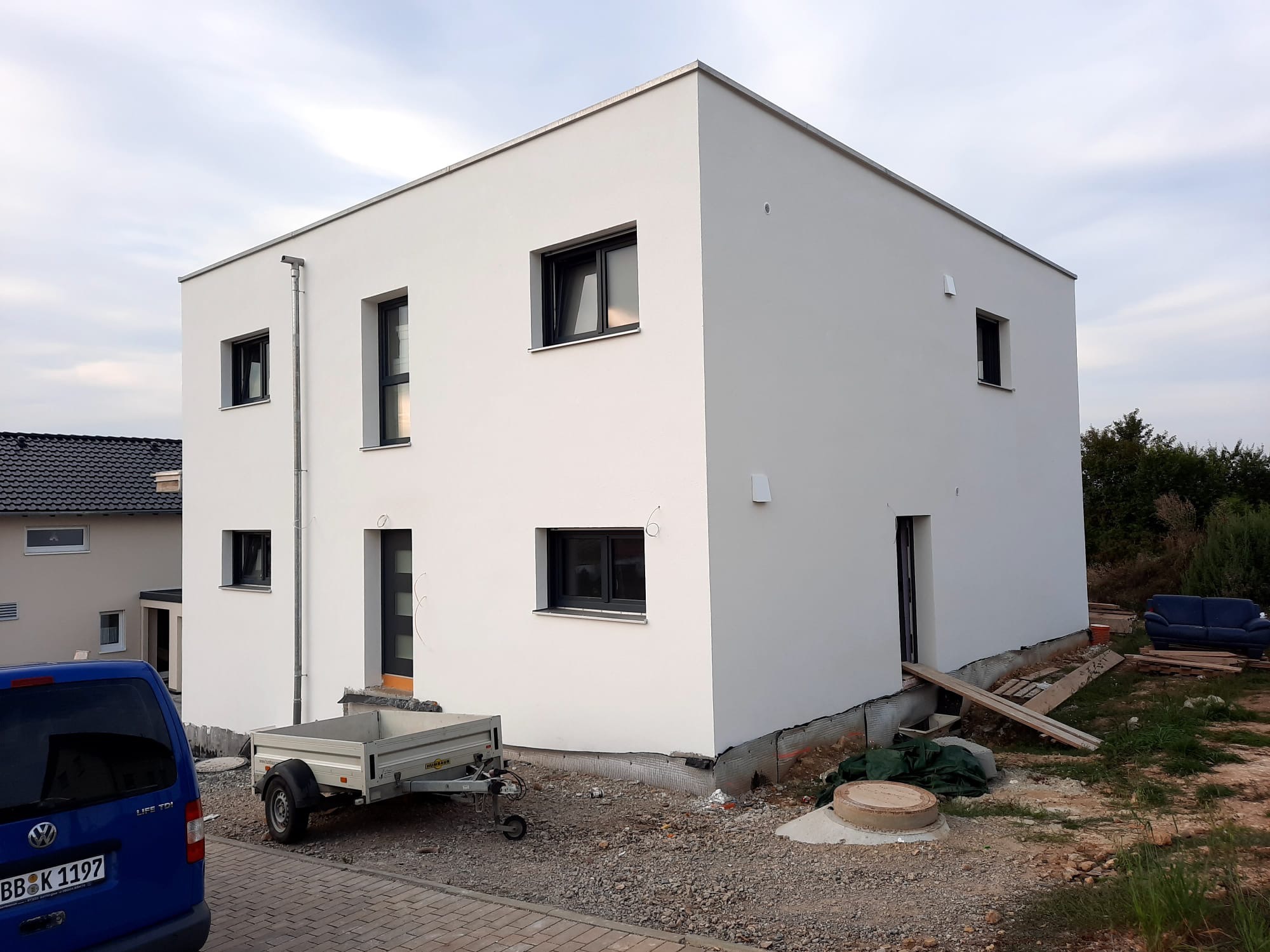 Nowoczesny, dwukondygnacyjny dom z białą elewacją, czarnymi oknami i przyczepką na posesji w trakcie prac wykończeniowych.