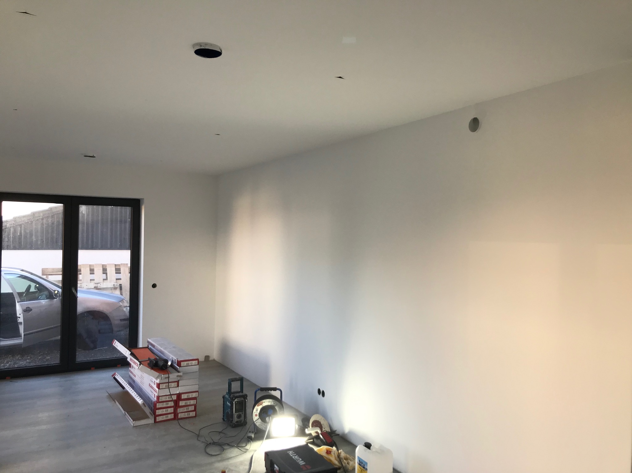 Wnętrze w trakcie prac wykończeniowych: pakiety paneli podłogowych, narzędzia oświetlone lampą, świeżo pomalowane ściany i widok na zewnątrz przez okno.