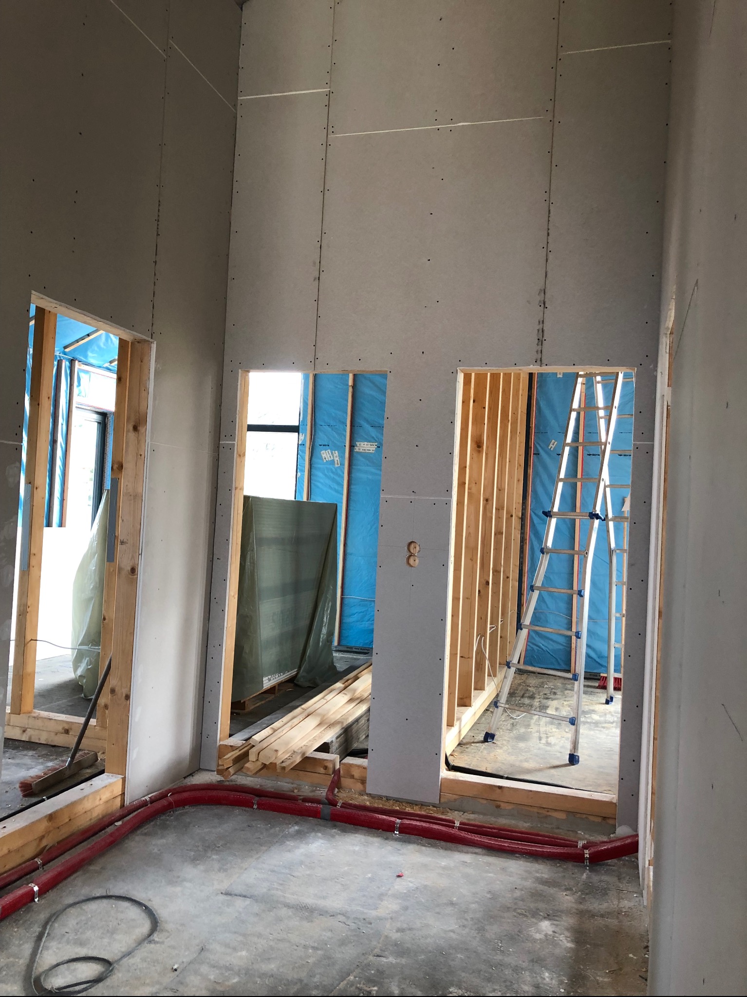 Wnętrze w trakcie wykańczania: ściany z płyt gipsowo-kartonowych, widoczne drewniane ramy otworów, instalacja podłogowa i drabina. Etap prac wykończeniowych.