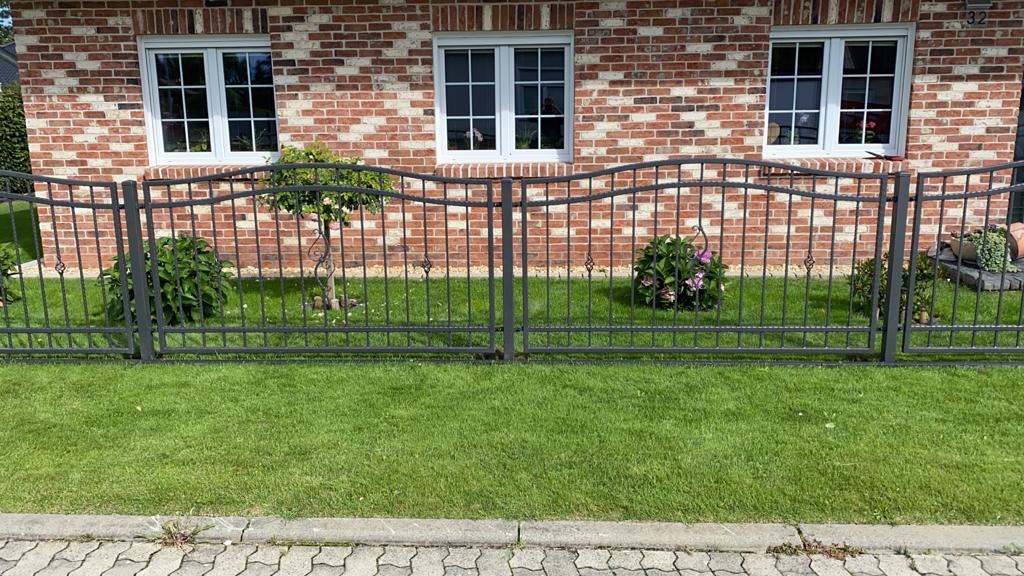 Metalowe ogrodzenie posesji z dekoracyjnymi elementami na tle ceglanego domu z trzema oknami, trawnik i roślinność wzdłuż ogrodzenia.