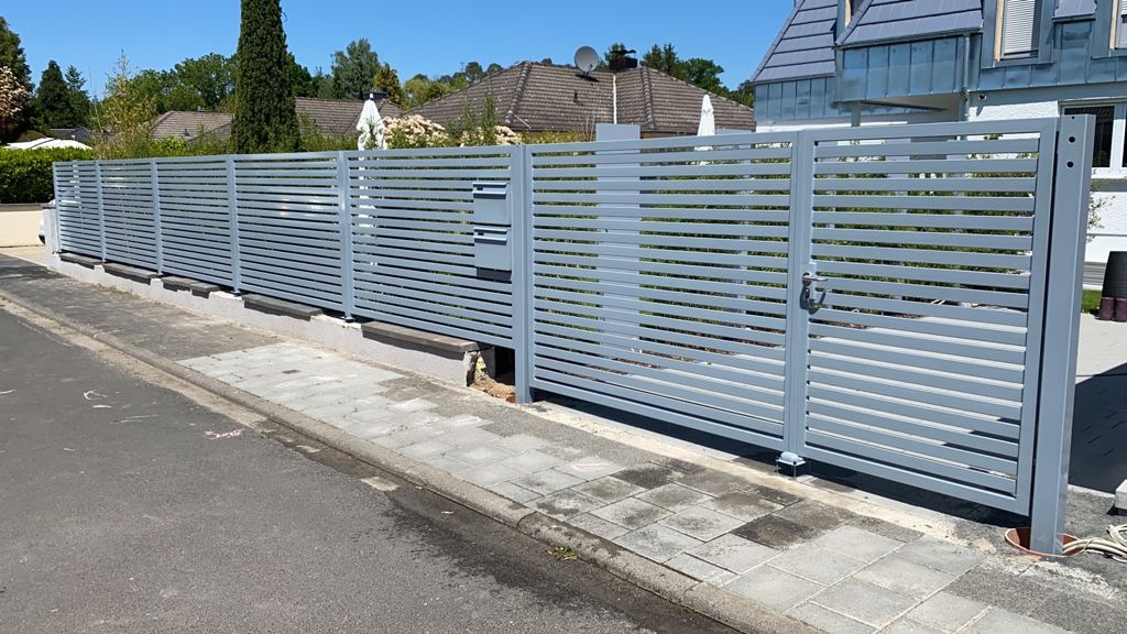Nowoczesne, szare ogrodzenie panelowe z poziomymi lamelami, z bramką wejściową, na tle zieleni i domów. Widok z poziomu chodnika, słoneczny dzień.