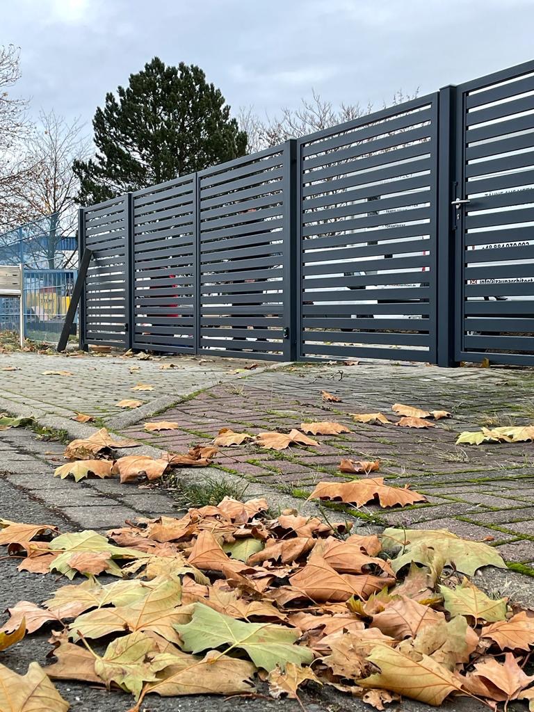 Nowoczesne ogrodzenie panelowe w kolorze grafitowym z poziomymi szczeblami, jesienne liście na brukowanym chodniku. Widok z dołu, pochmurne niebo.