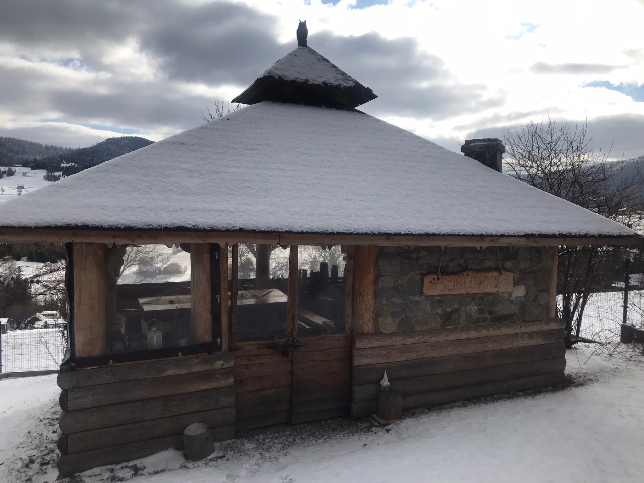 Zimowa altana z kamienia i drewna, pokryta śniegiem, z dekoracyjną figurką na dachu. W tle górski krajobraz i pochmurne niebo.