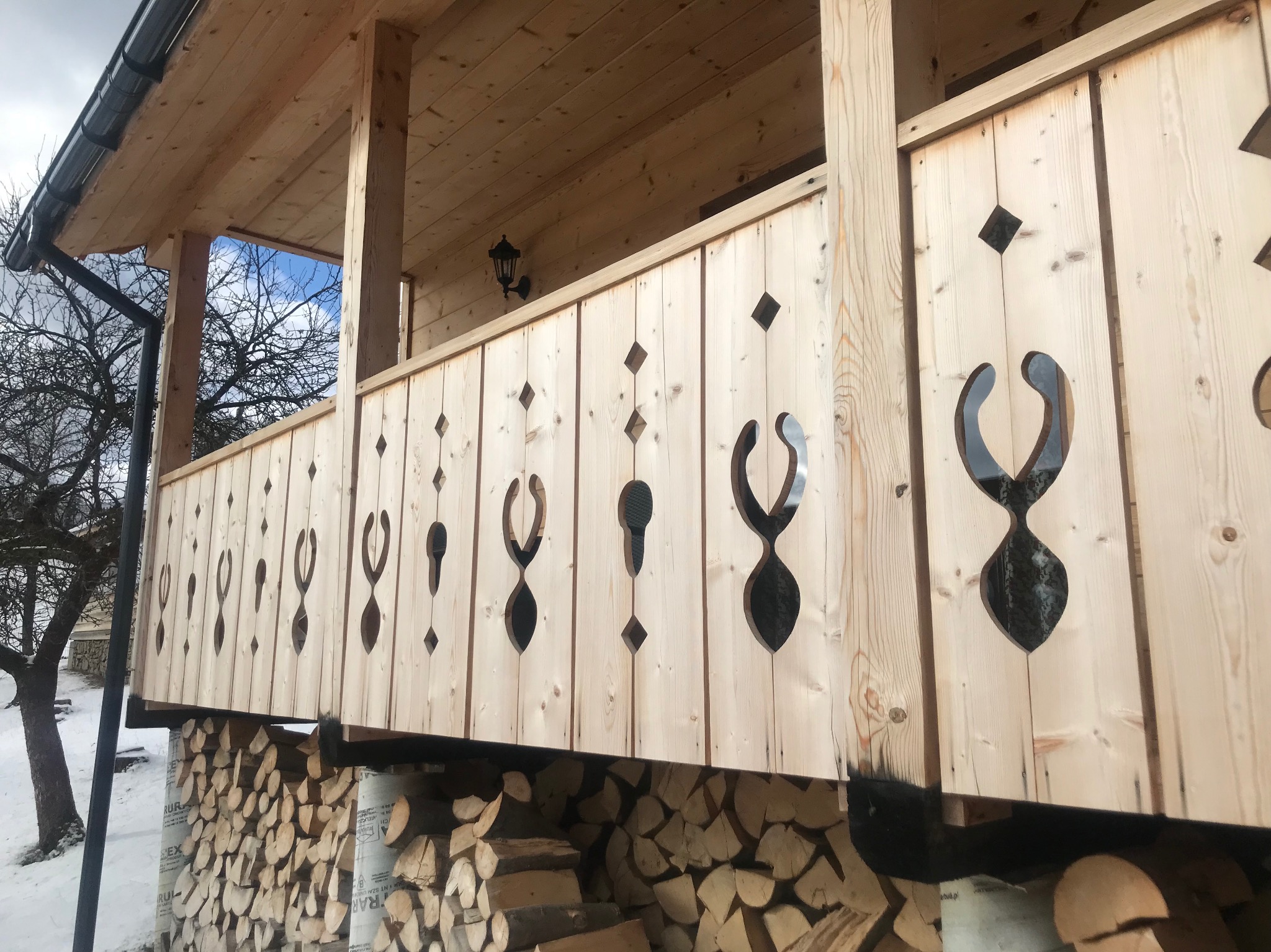 Drewniana pergola z dekoracyjnymi wycięciami, oświetlona lampą. Pod konstrukcją składowane drewno opałowe. Widok na zimowy krajobraz.