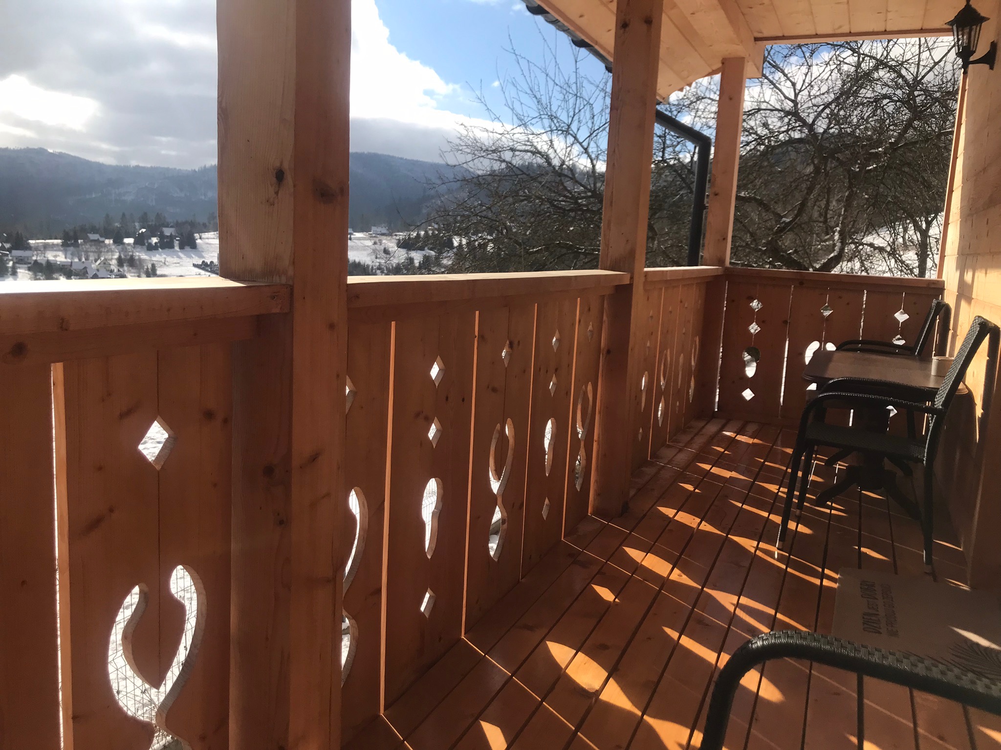Drewniany balkon z ażurową balustradą w górskim stylu, z widokiem na ośnieżone szczyty. Słońce rzuca geometryczne cienie na podłogę. W tle widać zabudowania.
