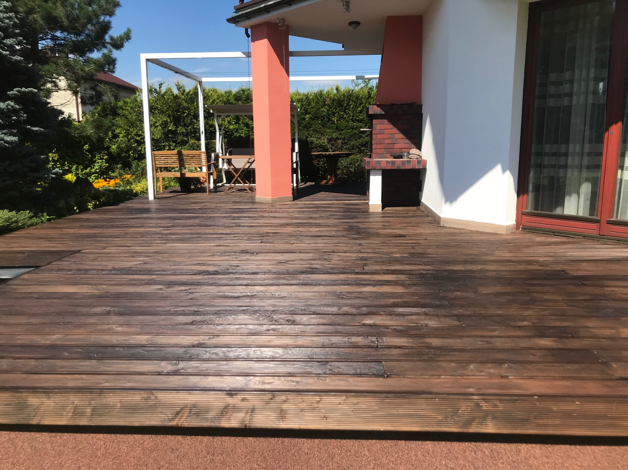 Ciemnobrązowy taras z desek w Soblówce, zadaszony pergolą, z grillem z cegły. Widok na ogród i dom.