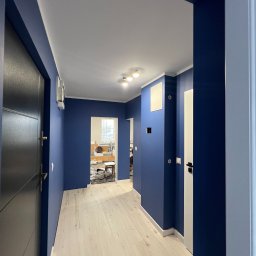 MATEUSZ SOWA SowRem - Korytarz po malowaniu na granatowo z jasną podłogą. Widoczne drzwi wejściowe i wewnętrzne, oświetlenie sufitowe. Ściany pomalowane na jednolity kolor, widoczne detale wykończeniowe.