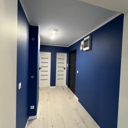 MATEUSZ SOWA SowRem - Korytarz po odświeżeniu: ściany w odcieniu głębokiego granatu, białe drzwi z poziomymi frezami, jasna podłoga imitująca drewno, minimalistyczne oświetlenie sufitowe.