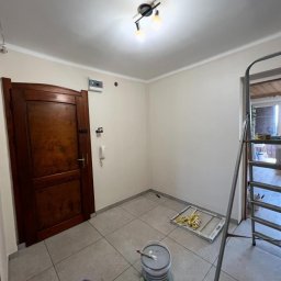 MATEUSZ SOWA SowRem - Wnętrze po malowaniu: jasne ściany, ciemne drzwi, drabina, wiadro z farbą i wałek na podłodze z szarych płytek. Widoczne gniazdka i włączniki na ścianie.