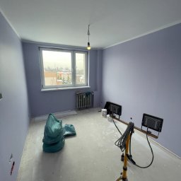 MATEUSZ SOWA SowRem - Jasny pokój po malowaniu ścian na fioletowo, widoczne worki z odpadami, lampa na statywie i wiadro z farbą. Grzejnik pod oknem. Świeżo pomalowane ściany.