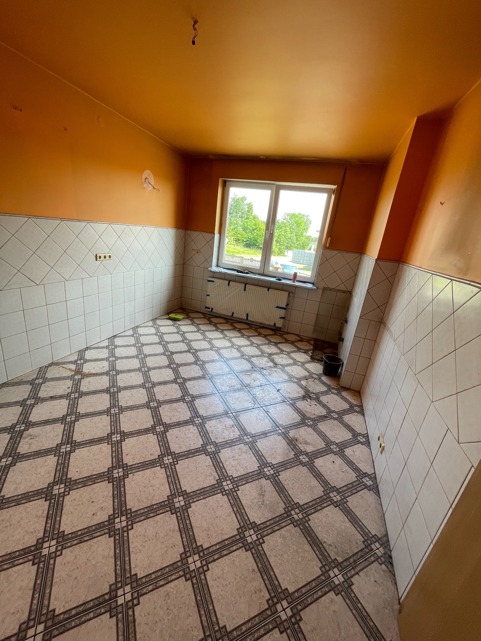 Pomarańczowa kuchnia przed remontem: stare płytki podłogowe i ścienne, okno z widokiem na zieleń. Widoczne ślady użytkowania i przygotowania do prac remontowych.